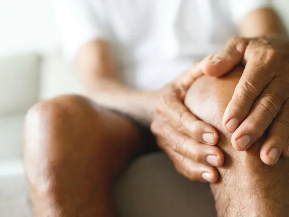 Seniorin hält sich beim Aufstehen aus dem Sessel das Knie – ein Hinweis auf die altersabhängige Gelenksteife und Anlaufschmerzen am Morgen.