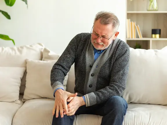 Ein älterer Mann sitzt auf einer Couch, hält sein Knie mit beiden Händen und scheint Schmerzen zu haben.